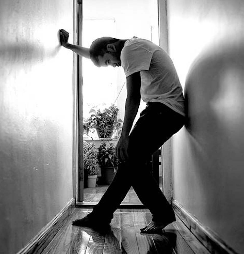 Black and white photo of young man standing up against a wall with his head slumped forward